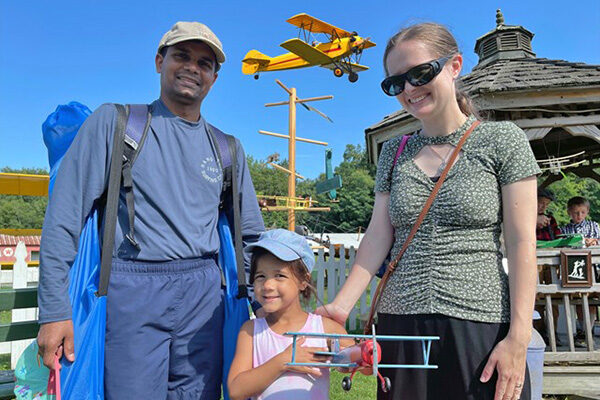 Photo of a family comprised of a father, mother, and child who is holding a top airplane. A yellow biplane is in mid-flight directly behind them.