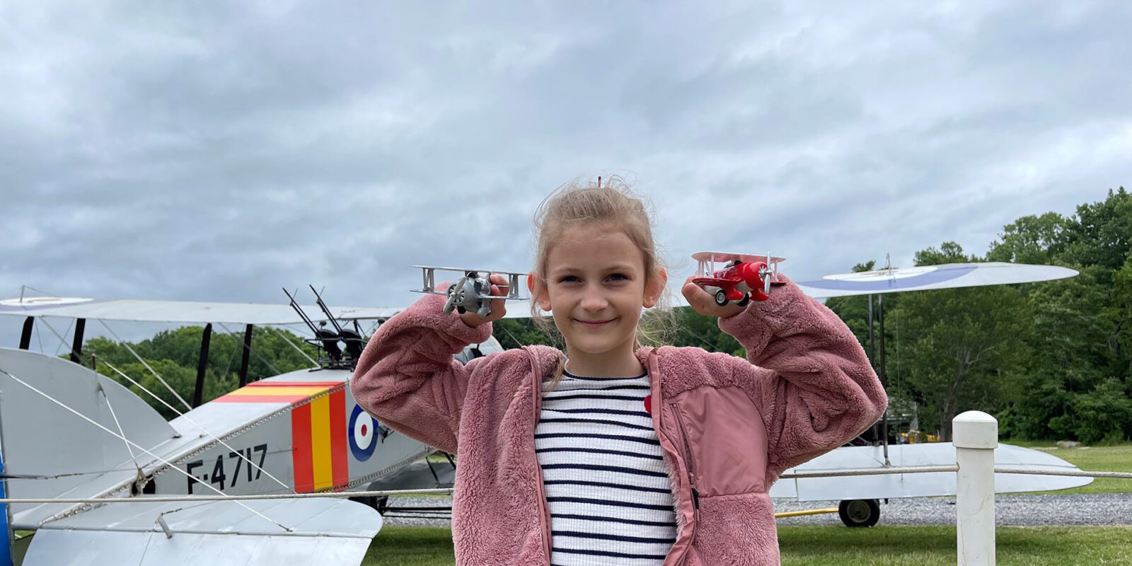 Photo of a girl holding up two model biplanes in front of an real biplane.