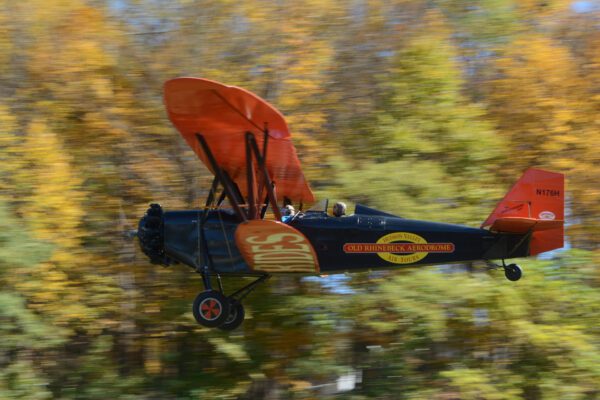 Photo of a blue and red biplane with "Old Rhinebeck Aerodrome" printed on the side racing across the screen from right to left in front blurred fall trees.